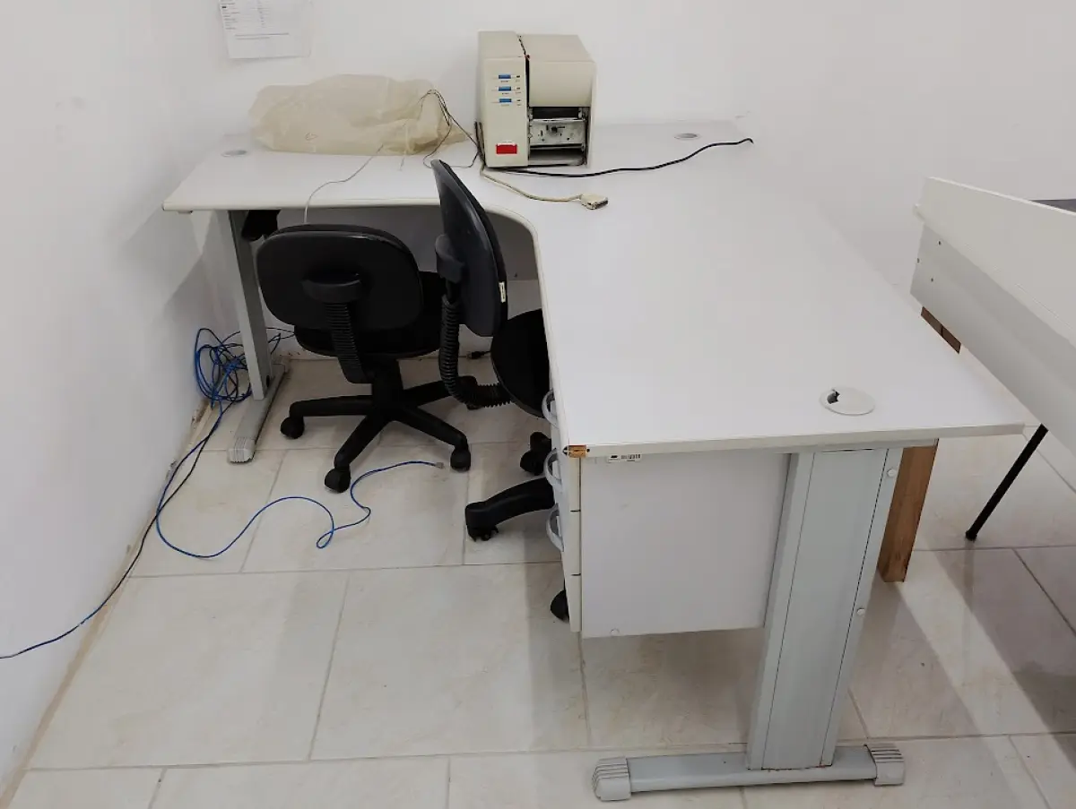 LOTE 132 - 01 mesa de escritório em "L" branca com 02 cadeiras, 01 equipamento (aparenta ser impressora/relógio ponto), 02 quadros brancos, 01 armário baixo de duas portas e 01 bancada/mesa de apoio branca.