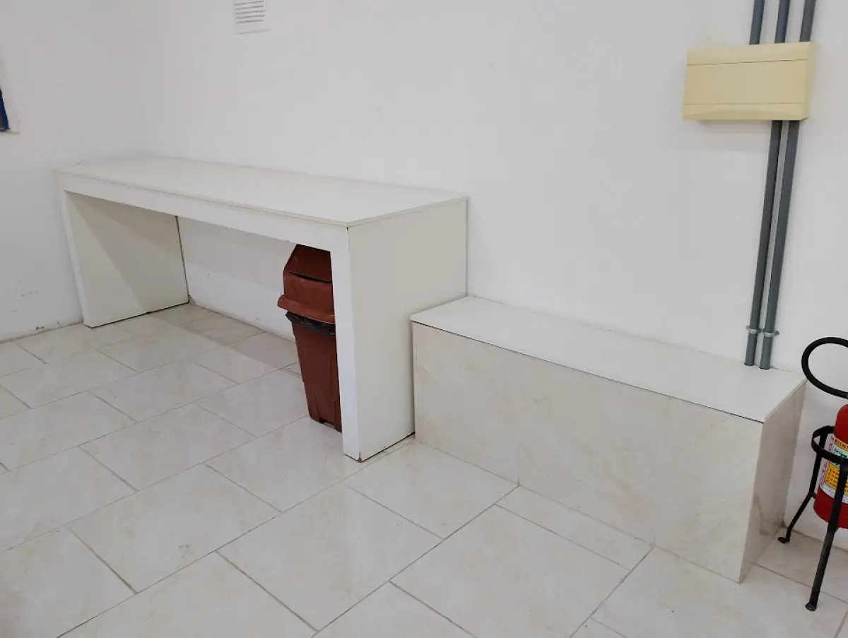 LOTE 132 - 01 mesa de escritório em "L" branca com 02 cadeiras, 01 equipamento (aparenta ser impressora/relógio ponto), 02 quadros brancos, 01 armário baixo de duas portas e 01 bancada/mesa de apoio branca.