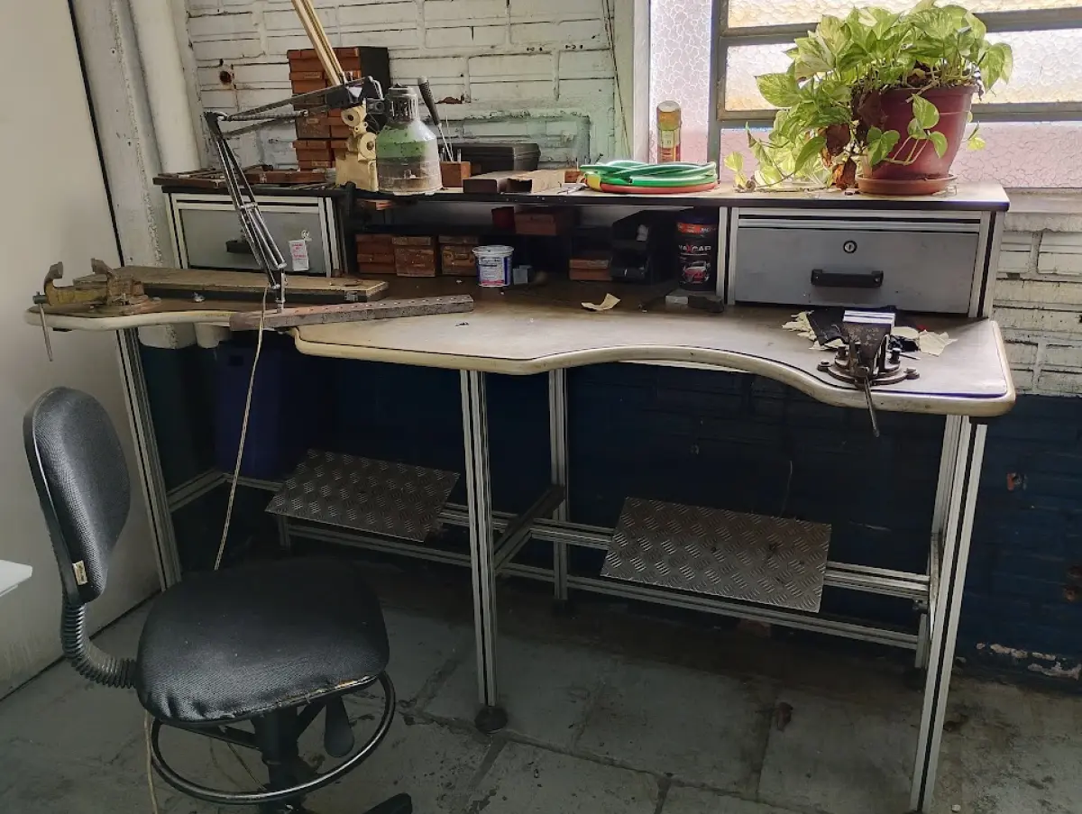 LOTE 111 - 01 bancada de trabalho ergonômica (estilo ferramenteiro/ourives) com estrutura em perfil de alumínio, tampos com recorte e apoios para os pés, 01 morsa de bancada marca Schulz nº 2, 01 morsa de bancada marca Modular nº 2 1/2, 01 luminária articulada, 01 cadeira giratória alta, pequeno lote de lixas, organizadores de madeira e miudezas em geral (nas gavetas).